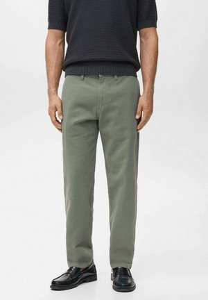 Mann trägt grüne gerade geschnittene Hose, schwarze Loafers und einen dunklen, strukturierten Kurzarmpullover, steht mit entspannten Armen an den Seiten.