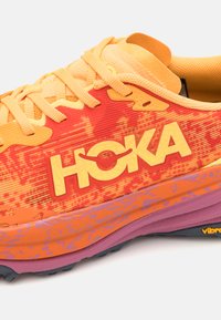 Chaussure de course Hoka en orange et rouge avec un dessus en maille texturée. Présente une semelle extérieure épaisse de couleur violette et un logo jaune bien visible sur le côté.