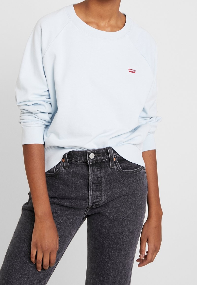 Personne portant un sweat Levi’s bleu clair avec un petit logo rouge sur la poitrine, associé à un jean en denim noir taille haute.