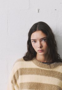 Pulover z valovitimi kremastimi in rjavimi črtami, z mehko teksturo, okroglim izrezom in prijetnim materialom, nosi oseba proti beli steni.