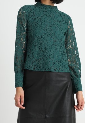 Femme portant un haut en dentelle florale vert foncé à manches longues, associé à une jupe en cuir noire sur un fond uni.