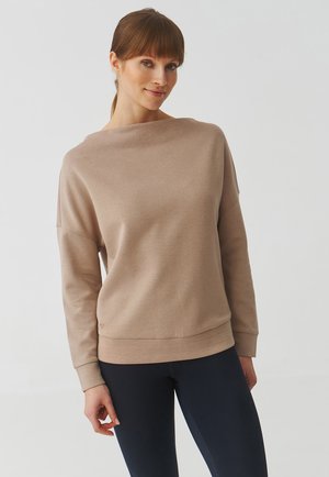 Felpa beige realizzata in tessuto morbido, con un ampio scollo a barca, spalle scese e orlo a coste, abbinata a leggings scuri.