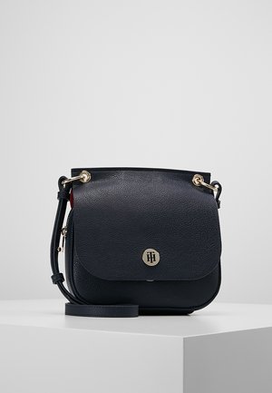 Sort tekstureret læder crossbody taske med klaplukning, guldfarvet hardware og justerbar rem, hvilende på en hvid overflade mod en grå baggrund.