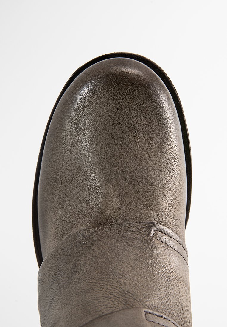 Gros plan sur l'orteil d'une botte en cuir gris usé, avec des coutures visibles et une surface texturée sur fond blanc.