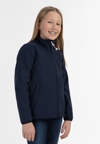 Jūrinio stiliaus zip-up striukė su aukštu apykakle, minkšta tekstūra, elastingais rankogaliais ir dviem šoninėmis kišenėmis, dėvima su mėlynomis džinsomis. Yra reguliuojama raištelių juosta.