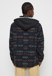 Hætte skjorte med knapper i mørkeblå med geometriske mønstre i brune og teal farver. Fleece materiale, struktureret med en kontrasterende sort hætte.