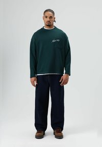 Grünes Langarmshirt mit einer Brusttasche und weißer Schrift, über einem weißen T-Shirt getragen, kombiniert mit dunkelblauen, übergroßen Jeans und Stiefeln.