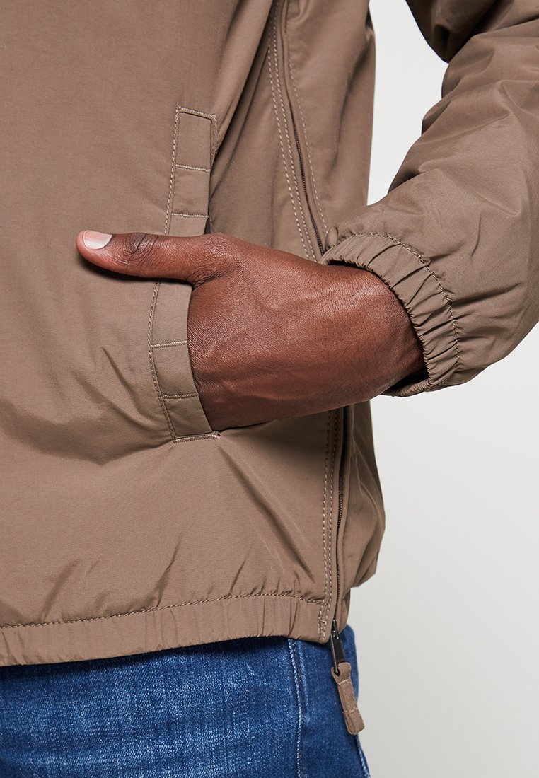 Chaqueta marrón con una textura suave, puños elásticos y bolsillos laterales. La tela parece ligera y presenta un cierre de cremallera.
