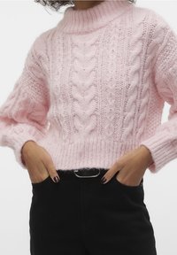 Personne portant un pull épais rose clair avec motifs torsadés, pantalon noir, mains dans les poches, présentant une tenue décontractée.