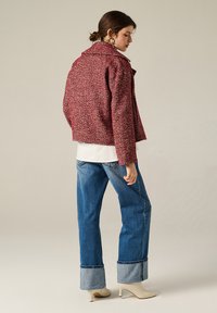 Giacca rossa con texture e motivo a spina di pesce, colletto a patta e spalle scese, abbinata a jeans in denim blu chiaro e décolleté crema.