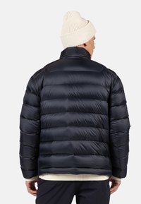 Giacca a vento blu navy con cuciture orizzontali, colletto alto e polsini a coste. Indossata sopra un maglione di colore chiaro e abbinata a un cappello di maglia.