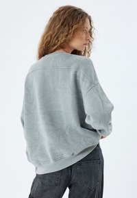 Felpa verde sbiadita con vestibilità comoda, scollo rotondo e maniche raglan. Tessuto testurizzato e orlo a costine, abbinata a jeans scuri.
