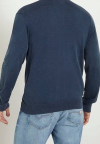 Mann in einem dunkelblauen Langarmhemd und hellblauen Jeans, von hinten vor einem schlichten Hintergrund gezeigt.