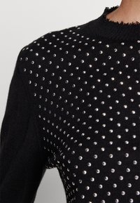 Pull en maille noire avec de petits clous argentés brillants couvrant uniformément le haut de la poitrine et les épaules, montrant un détail de tissu texturé.