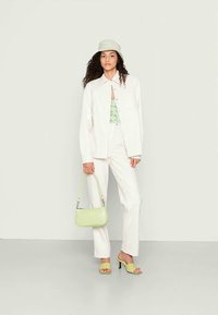 Camisa branca de botão, top de alças com padrão verde, calças brancas, bolsa verde, sandálias de salto alto verdes, cabelo encaracolado, fundo neutro.