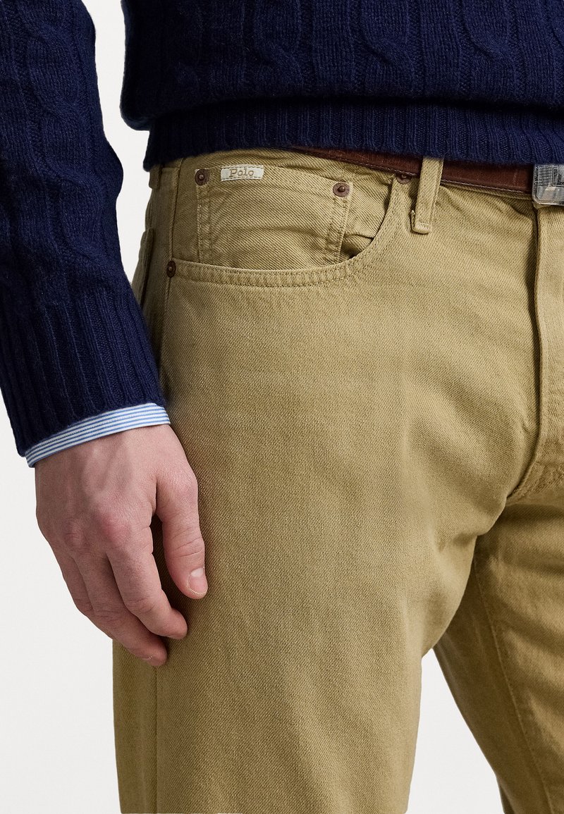 Khakifarbene Baumwollhose mit klassischer Passform, mit Fronttaschen, einem braunen Ledergürtel und einem blauen Kabelstrickpullover mit gestreiftem Kragen.