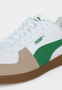 Baskets blanches Puma avec bande latérale verte, détails en daim beige, lacets blancs et semelle en gomme marron.