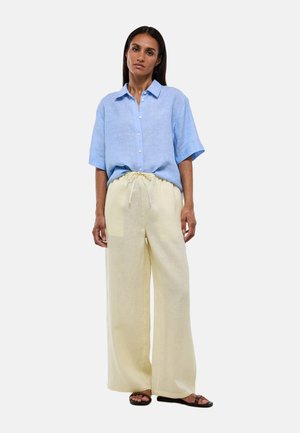 Femme portant une chemise à manches courtes bleu clair à boutons, un pantalon ample taille haute jaune clair et des sandales noires, debout sur un fond uni.
