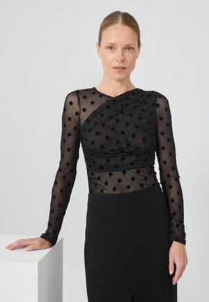 Haut noir à manches longues en tissu transparent à pois, présentant un design drapé et une silhouette ajustée, associé à une jupe noire unie.