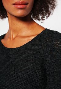 Svart, stickad topp med vid halsringning, strukturerat tyg och en något lös passform. Sömmen skapar ett vågmönster över hela plagget.