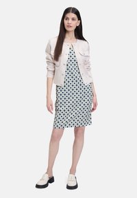 Helles beiges Jackett mit Knopfverschluss über einem enganliegenden Kleid mit einem geometrischen Blau- und Grünton. Das Model trägt cremefarbene Loafers.