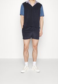 Polo en tissu éponge navy à manches courtes avec col violet clair, associé à un short assorti. Baskets blanches avec œillets colorés et chaussettes montantes.