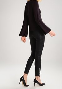 Kvinna som går i en svart topp med utsvängda ärmar, figurnära svarta byxor med blixtlås vid anklarna och svarta högklackade skor mot en ljus bakgrund.