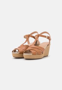 Sandales compensées en cuir tan avec des brides croisées, design à bout ouvert et plateforme en texture tissée. Accent en boucle dorée. Semelle intérieure légèrement rembourrée.