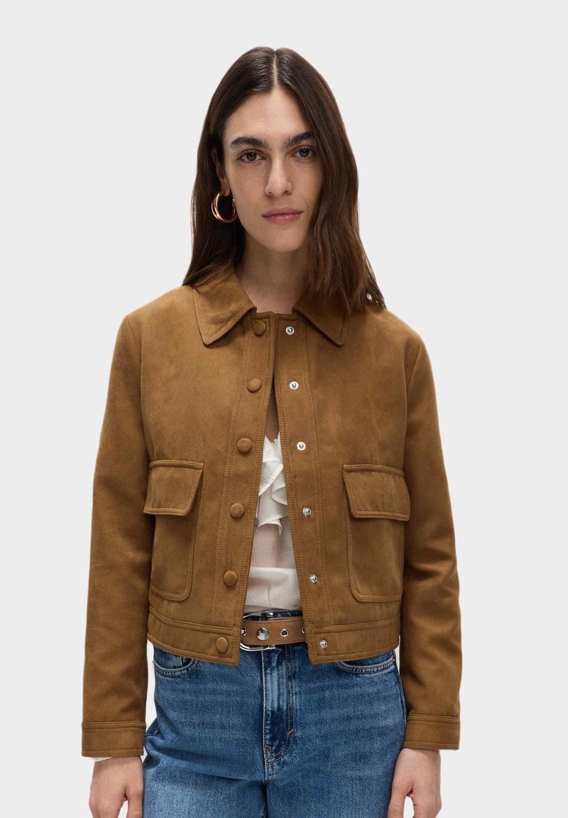 Giacca in suede marrone con colletto, tasche frontali e chiusura a bottone a pressione, abbinata a una blusa bianca con balze e jeans in denim.