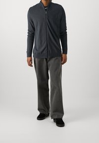 Mörkgrå zip-up-tröja med långa ärmar, tillverkad av ett slätt tyg, kombinerad med löst sittande grå byxor och svart fotbeklädnad.