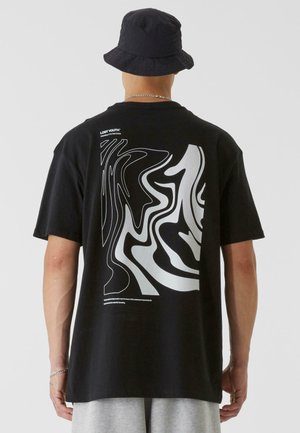 Homme portant un bob noir et un T-shirt noir avec un motif blanc abstrait ondulé et du texte au dos, assorti à un short gris clair.