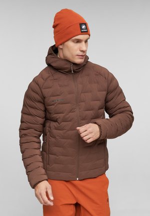 Mann trägt braune gesteppte Jacke, orange Mütze mit Mammut-Logo und orange Hose, schaut nach rechts vor neutralem Hintergrund.