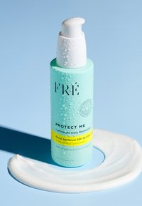 Miętowozielona butelka z białą pompką, oznaczona napisem "Chroń mnie." Teksturowana kroplami, leżąca obok wiru białej krema na jasnoniebieskiej powierzchni.