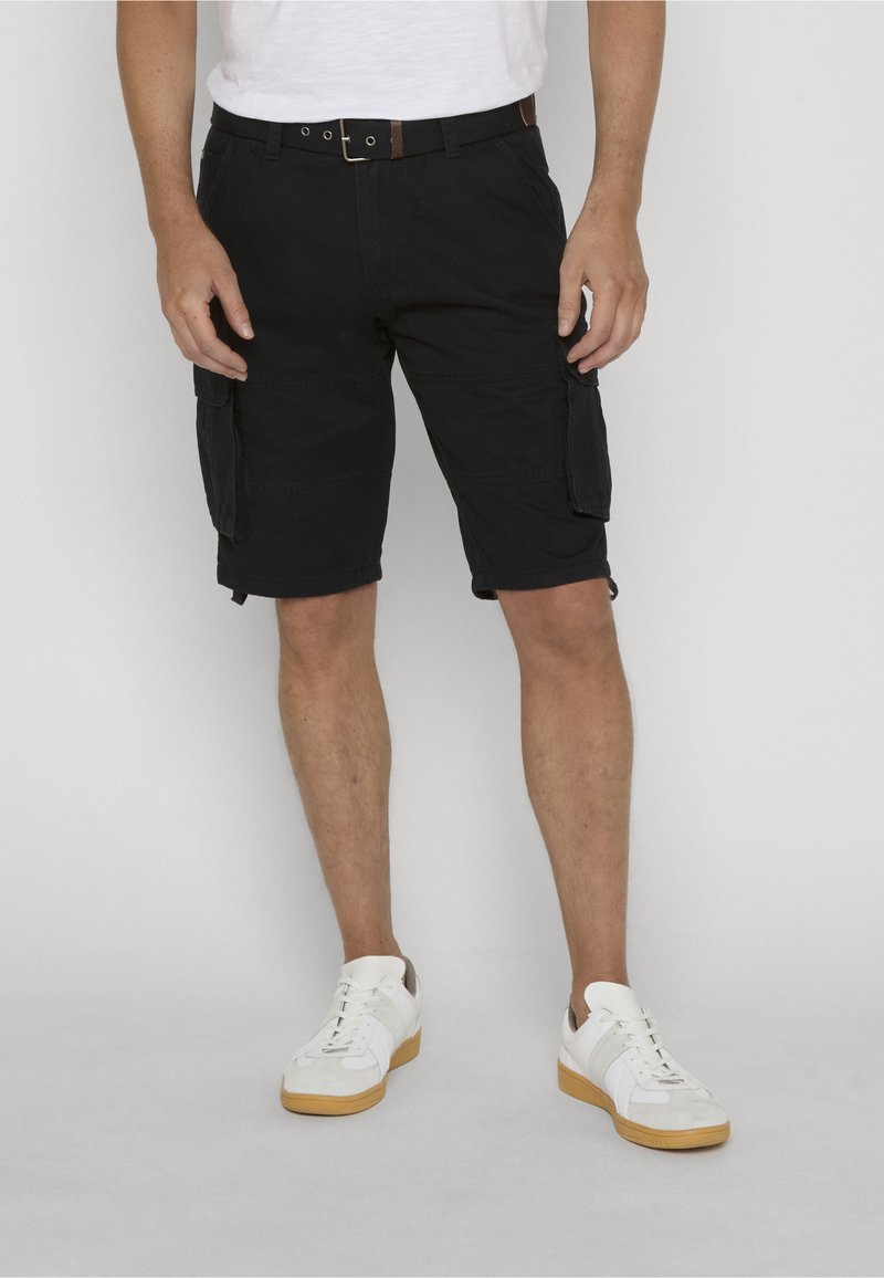 Homme portant un short cargo noir, une ceinture blanche, des baskets blanches, debout sur un fond clair.