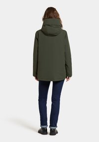 Groene parka met hoge kraag, capuchon en rechte snit, gedragen met donkere jeans en zwarte schoenen. Glad stofoppervlak.