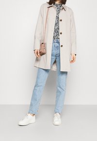 Beige trenchcoat med bruna knappar, blommig skjorta, ljusblå jeans och vita sneakers. En liten rosa axelväska hänger på axeln.