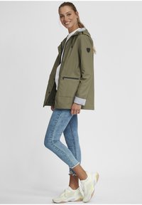 Veste imperméable vert olive avec capuche, fermeture éclair à l'avant et deux poches latérales. Associée à un jean bleu déchiré et des baskets blanches.
