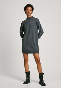 Abito di maglia grigio con scollatura alta e orlo a coste, abbinato a stivali neri alla caviglia, su uno sfondo neutro.