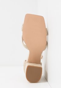 Sandale à talon beige avec bout carré et talon bloc texturé, positionnée semelle vers le haut contre un coin de mur blanc.