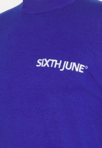 Blå högkrage tröja gjord av ett slätt stickat tyg, med det vita broderade varumärkesnamnet "SIXTH JUNE" på bröstet.