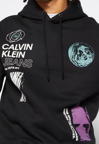 Czarna bluza z kapturem z białymi i turkusowymi grafikami, w tym tekstem "Calvin Klein Jeans" oraz abstrakcyjnymi wzorami w kolorze purpurowym i białym.