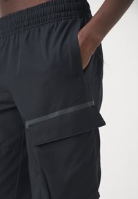 Czarne spodnie cargo z elastycznym pasem, bocznymi kieszeniami oraz detalem kieszeni na klapę na udzie. Gładka, lekka tkanina.