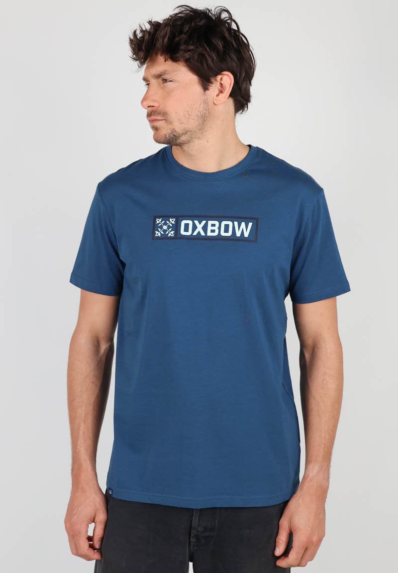 T-shirt bleu à manches courtes en coton. Dispose d'un graphique rectangulaire avec du texte "OXBOW" et des motifs de flocons de neige sur le devant.