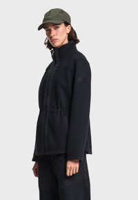 Femme portant une veste noire à fermeture éclair et une casquette vert olive, faisant face avec une expression neutre sur un fond uni.