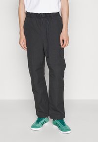 Persoon die zwarte cargobroek met verstelbare riem en blauwe sneakers draagt, staand tegen een effen witte achtergrond.