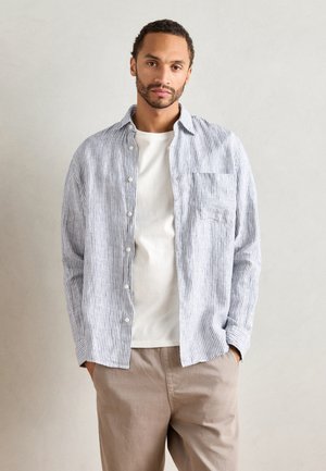 Button-up shirt featuring blue and white stripes, long sleeves, and a chest pocket, paired with a plain white T-shirt and beige pants.