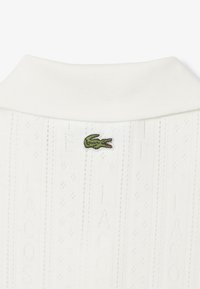 Polo en maille blanche avec un col côtelé, présentant un motif texturé et un petit logo de crocodile brodé vert à l'arrière.