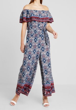 Femme portant une combinaison à épaules dénudées et jambes larges avec un motif cachemire bleu, rouge et blanc, ainsi que des chaussures blanches à bout ouvert.