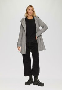 Manteau gris à capuche avec fermeture éclaire et boutons, associé à un pull côtelé noir et un pantalon large en velours côtelé noir. Bottes noires à semelle épaisse.