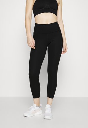 Leggings noirs taille haute avec une texture lisse et un matériau élastique. Associés à un soutien-gorge de sport noir court et à des baskets blanches.